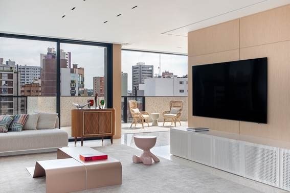 Sala de estar com painel de madeira clara, sofá cinza e banco Toy em rosa ao lado de mesa de centro baixa em tom blush, com varanda e vista da cidade ao fundo