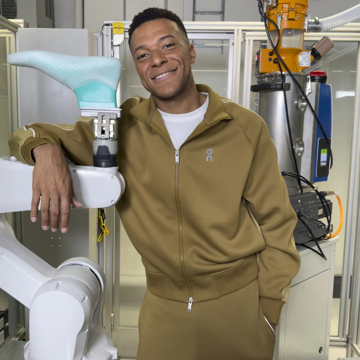 Kylian Mbappé sorri com conjunto esportivo verde-oliva da On, apoiado em um braço robótico que segura uma forma de calçado em um laboratório industrial