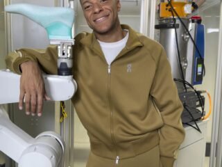 Kylian Mbappé sorri com conjunto esportivo verde-oliva da On, apoiado em um braço robótico que segura uma forma de calçado em um laboratório industrial