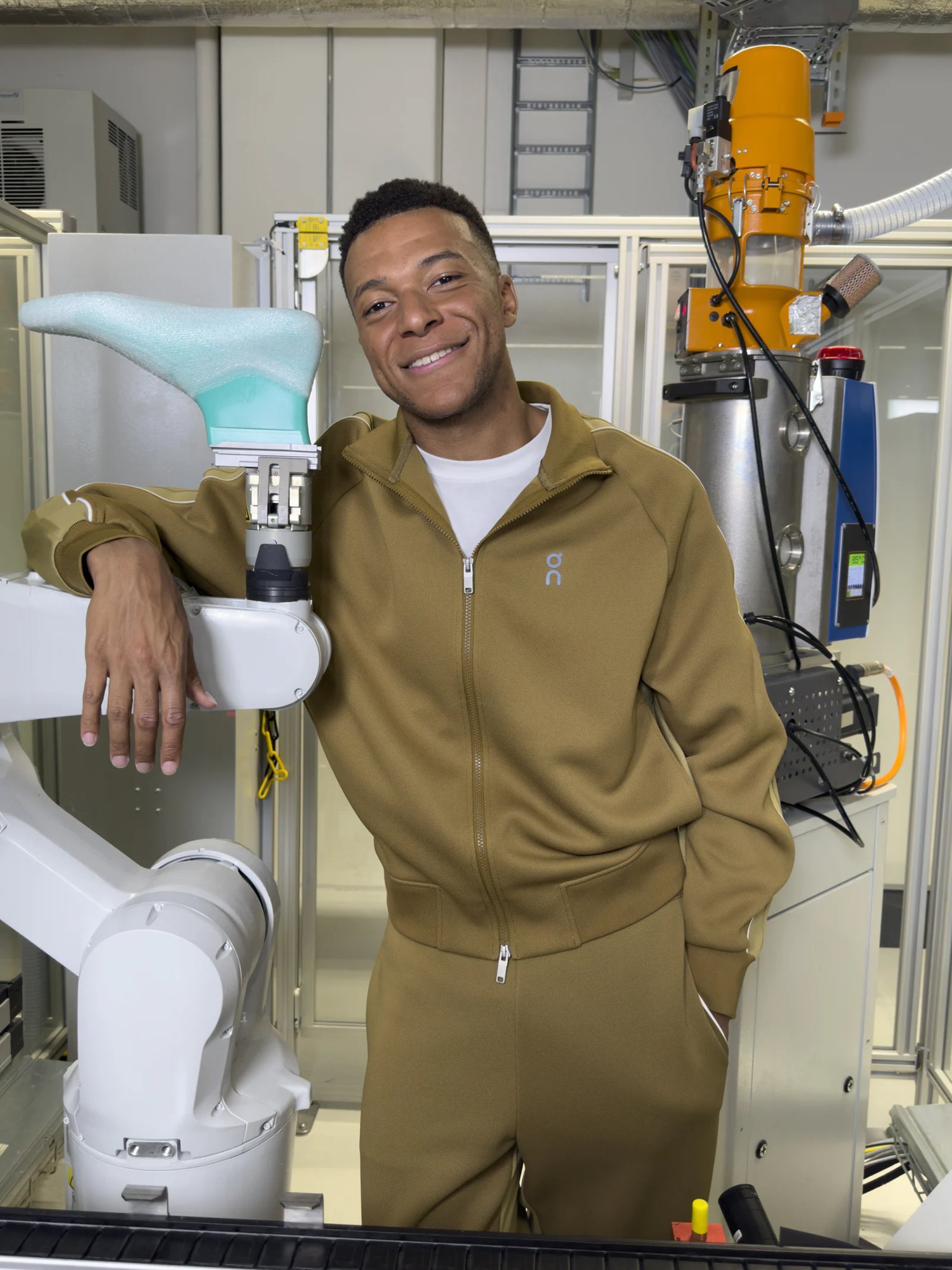 Kylian Mbappé sorri com conjunto esportivo verde-oliva da On, apoiado em um braço robótico que segura uma forma de calçado em um laboratório industrial