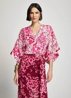 Modelo de cabelo curto usa blusa transpassada de estampa floral em rosa e vinho, com mangas amplas estilo kimono, sobre saia da mesma estampa