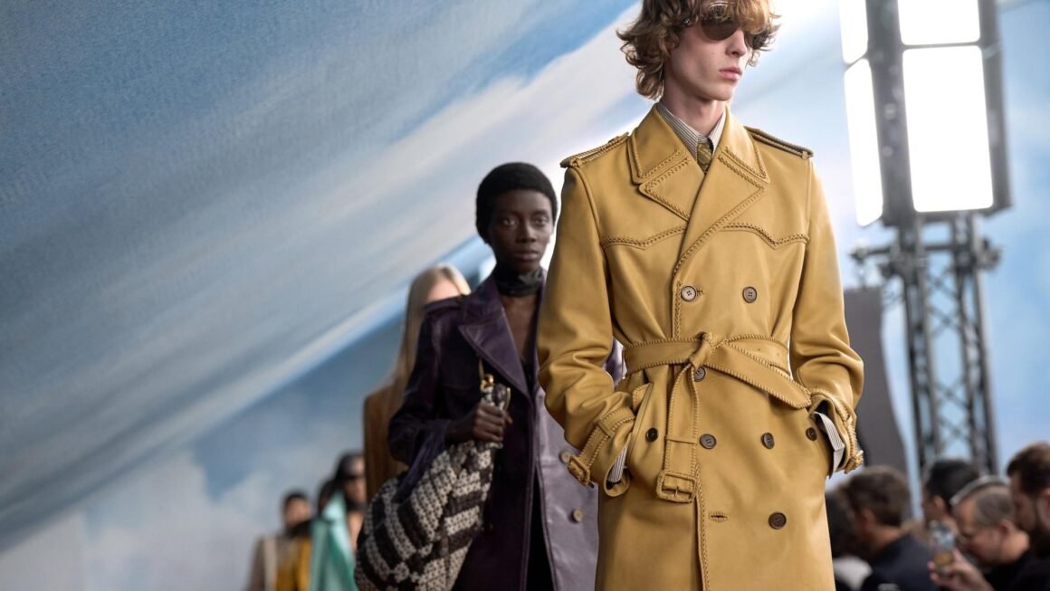Modelo de óculos escuros e trench coat de couro mostarda com cinto desfila à frente de outros modelos na passarela, com cenário de céu azul ao fundo