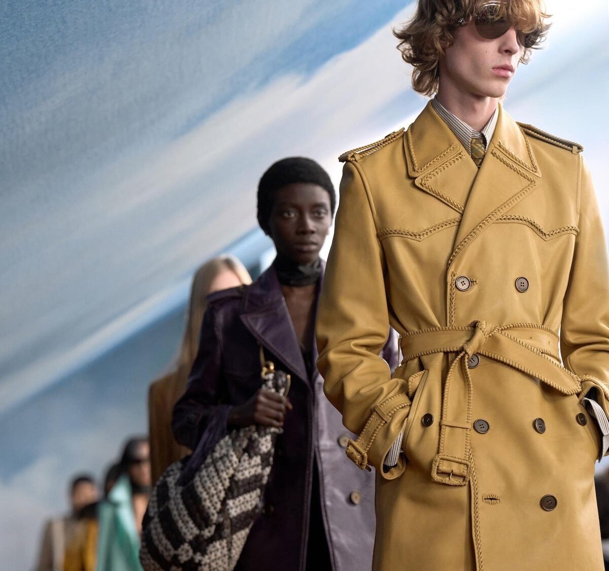 Modelo de óculos escuros e trench coat de couro mostarda com cinto desfila à frente de outros modelos na passarela, com cenário de céu azul ao fundo