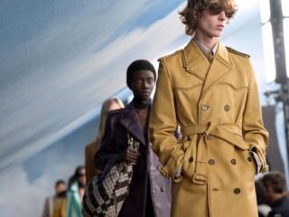 Modelo de óculos escuros e trench coat de couro mostarda com cinto desfila à frente de outros modelos na passarela, com cenário de céu azul ao fundo