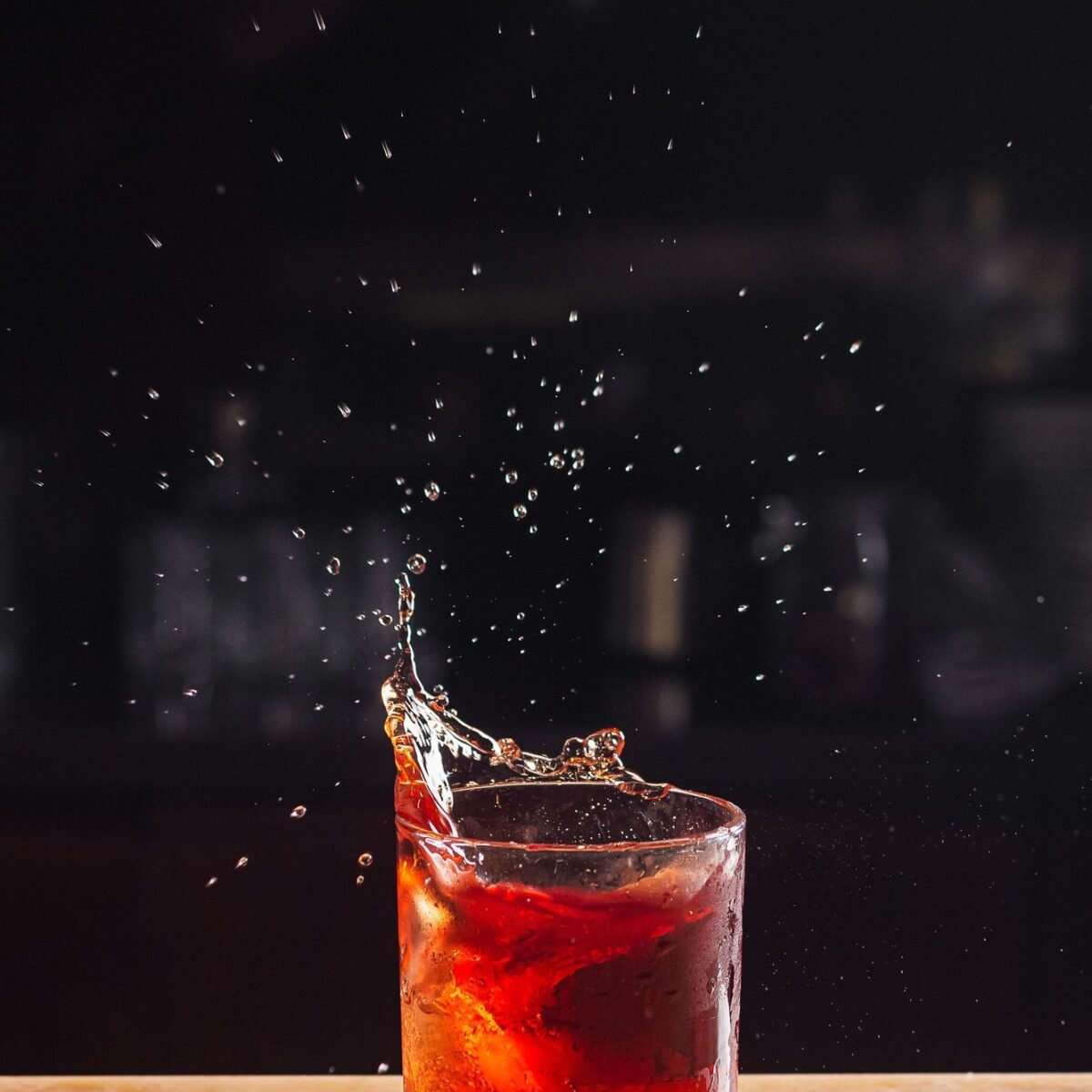 Copo baixo com drinque vermelho e gelo sobre balcão de bar, com respingos suspensos no ar e fundo escuro
