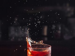 Copo baixo com drinque vermelho e gelo sobre balcão de bar, com respingos suspensos no ar e fundo escuro