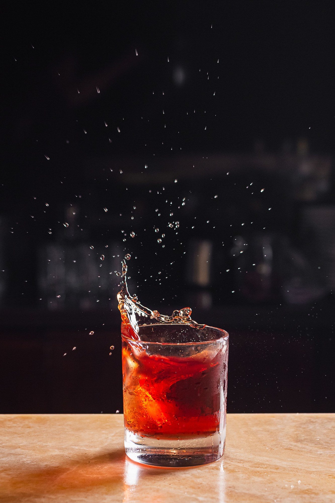 Copo baixo com drinque vermelho e gelo sobre balcão de bar, com respingos suspensos no ar e fundo escuro