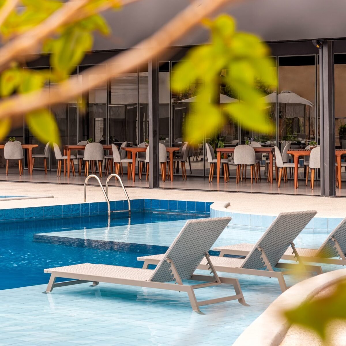 Três espreguiçadeiras dentro da borda rasa da piscina do Tivoli Maiorana, com o restaurante envidraçado ao fundo e folhagem em primeiro plano