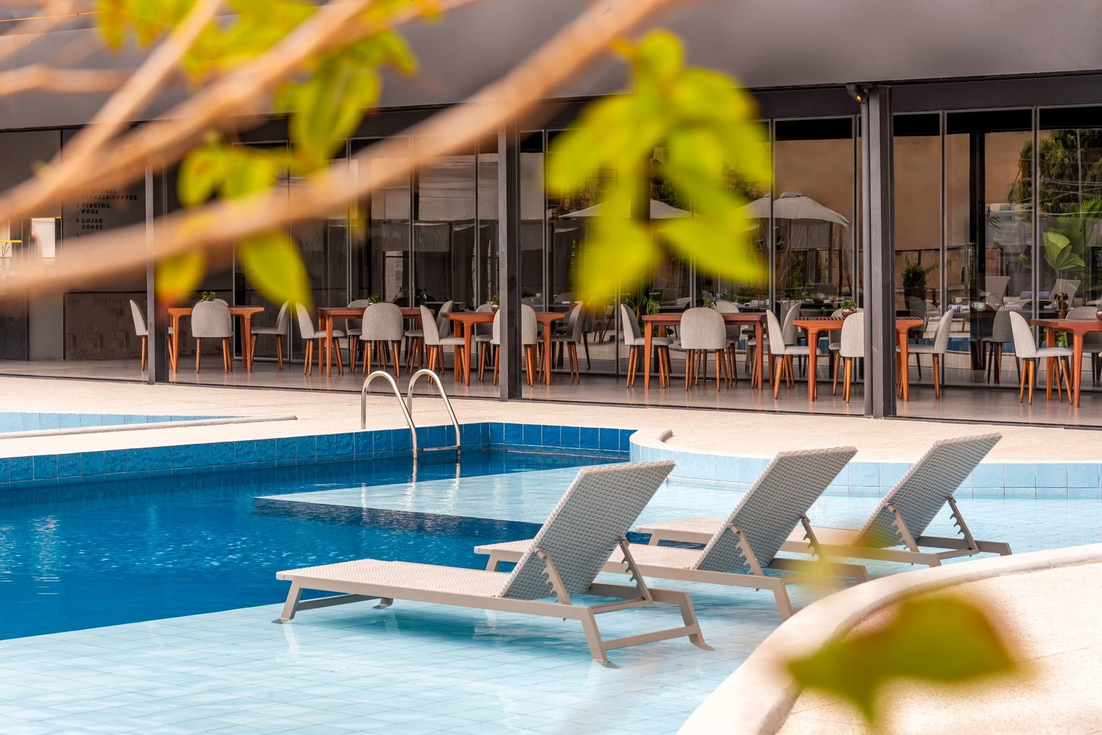 Três espreguiçadeiras dentro da borda rasa da piscina do Tivoli Maiorana, com o restaurante envidraçado ao fundo e folhagem em primeiro plano