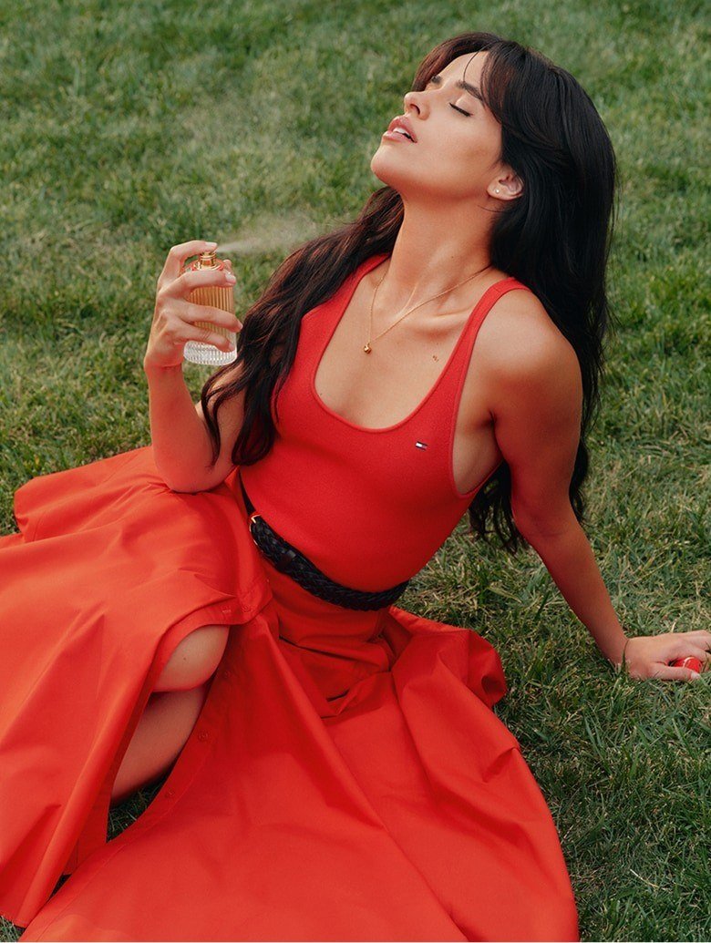 Camila Cabello deitada na grama, de vestido vermelho, borrifando um frasco de perfume dourado