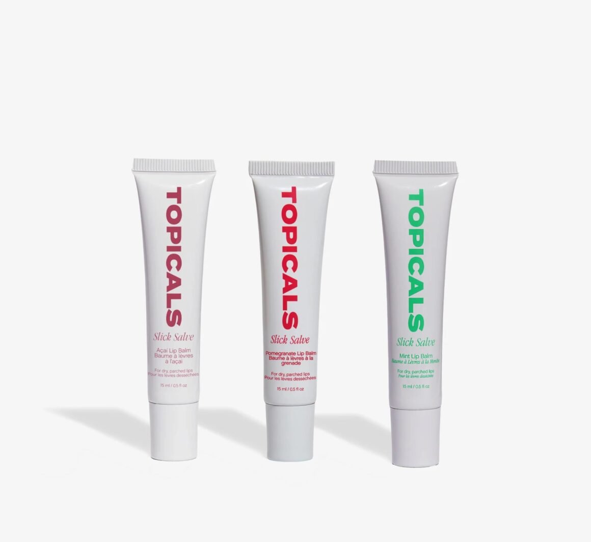 Três bisnagas brancas do lip balm Slick Salve da Topicals, nas versões açaí, romã e menta, em fundo claro