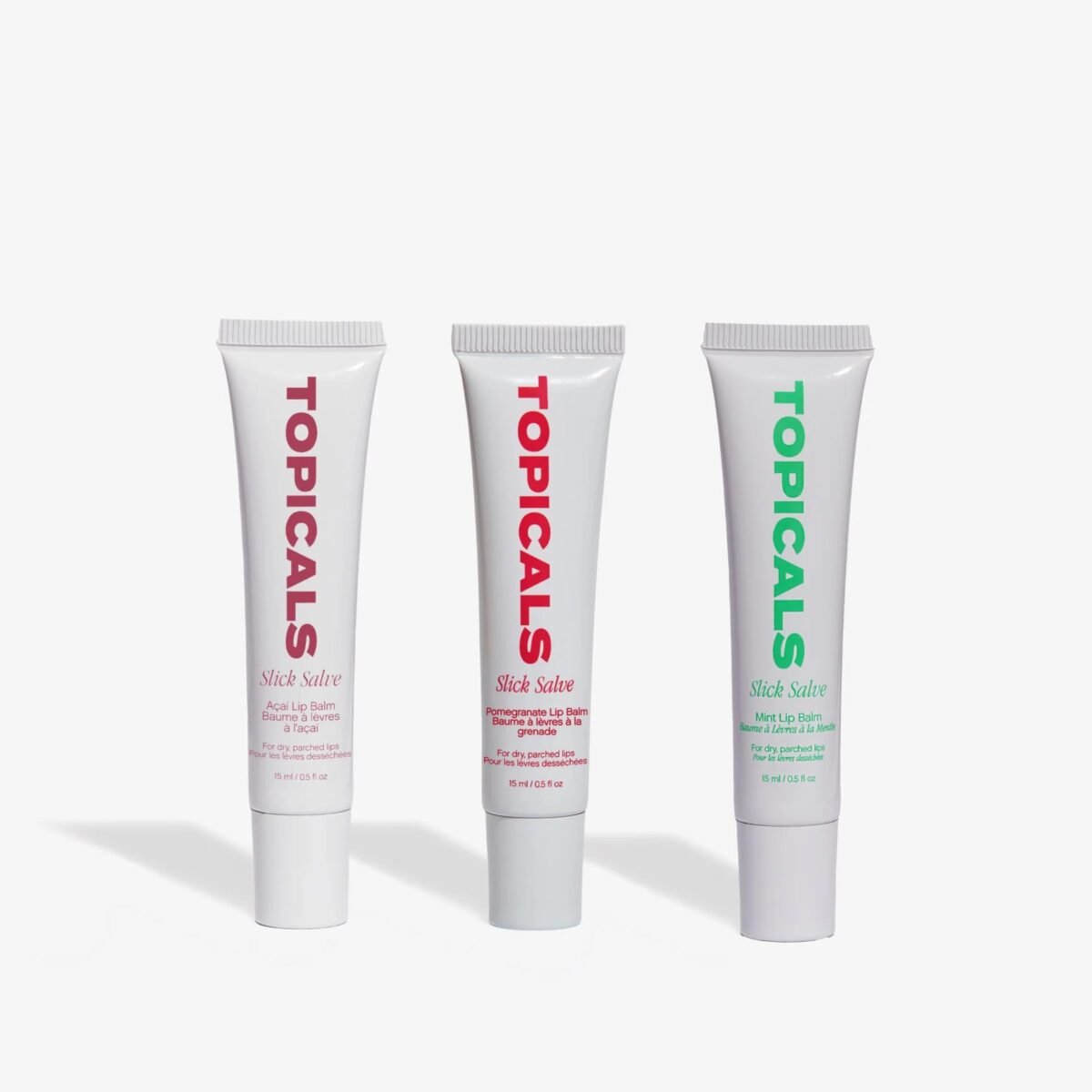 Três bisnagas brancas do lip balm Slick Salve da Topicals, nas versões açaí, romã e menta, em fundo claro