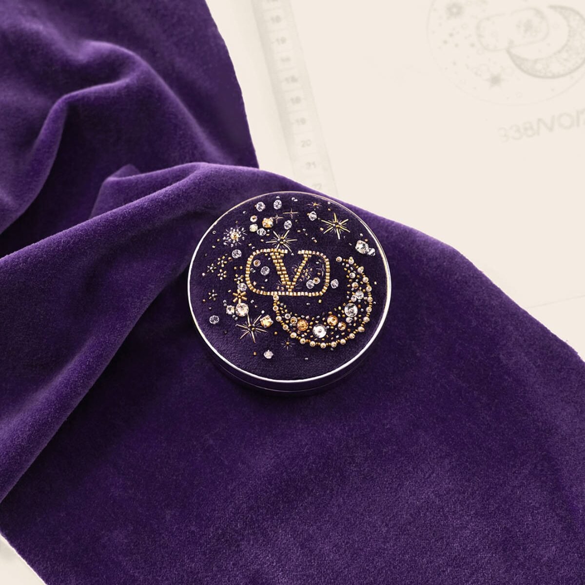 Compacto roxo bordado com motivo celeste sobre tecido de veludo.