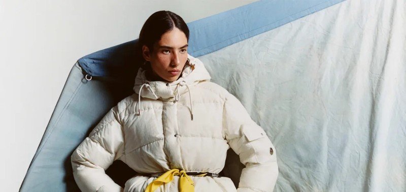 Modelo veste jaqueta acolchoada branca da coleção Moncler Re/Icons 2026, com faixa amarela na cintura.