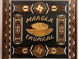 Quadro "Marola Tropical", de Thiago Nevs, em marchetaria de madeiras com moldura ornamentada, letreiro dourado e uma paisagem de praia com coqueiro e sol ao centro