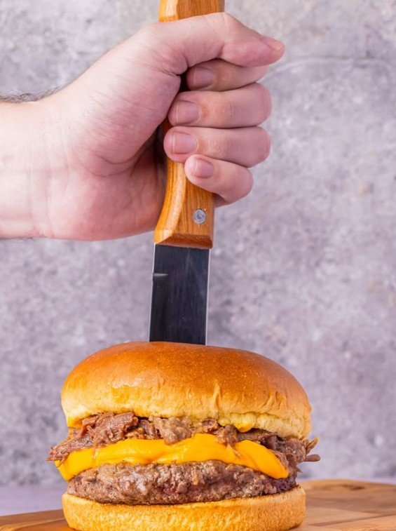 Hambúrguer com carne desfiada e queijo cheddar em pão brioche, com uma faca cravada no topo segurada por uma mão, sobre tábua de madeira