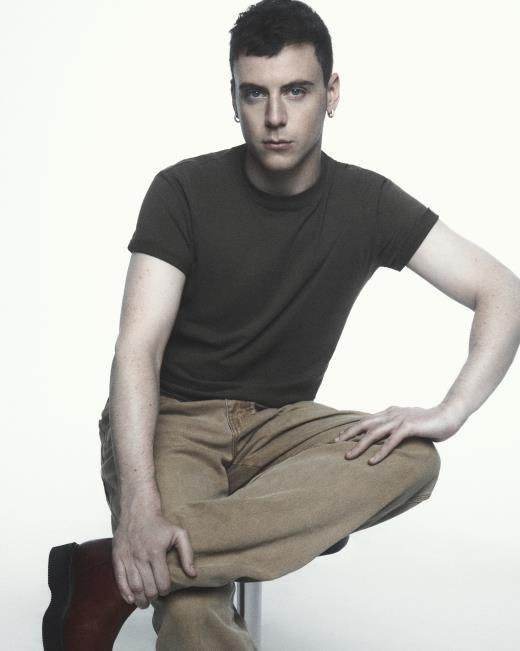 Retrato de Seán McGirr com camiseta marrom e calça cáqui, sentado sobre fundo branco