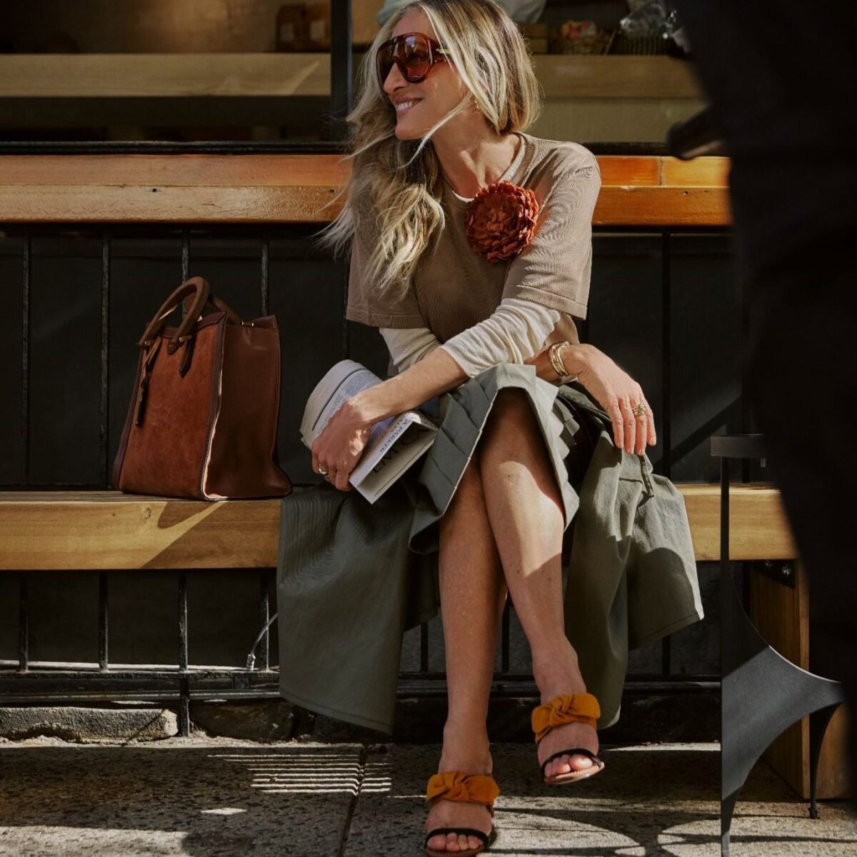 Sarah Jessica Parker sentada em um banco em Nova York, com sandália de laço em camurça, bolsa de camurça e couro e óculos grandes, na campanha Verão 27 da Arezzo