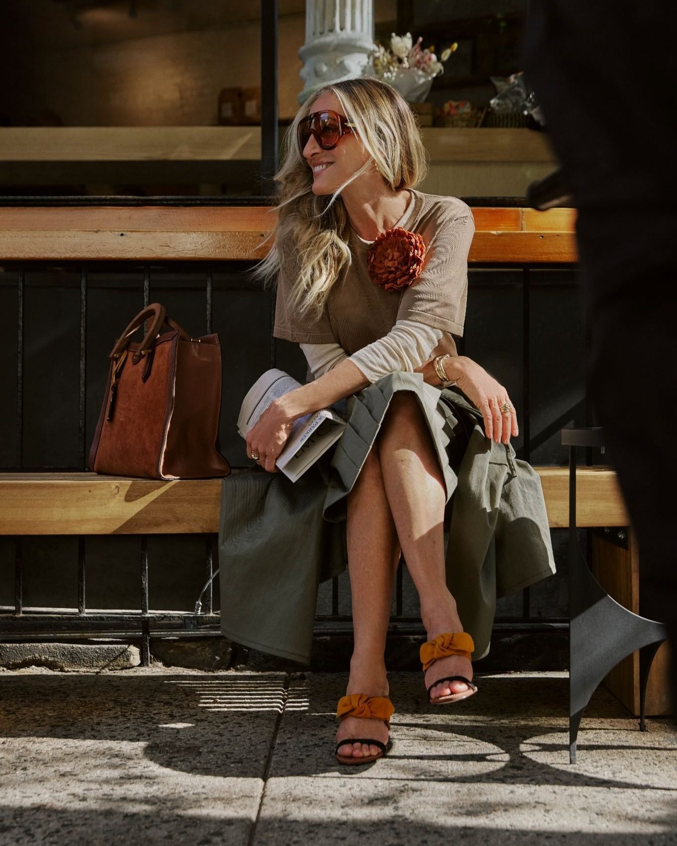 Sarah Jessica Parker sentada em um banco em Nova York, com sandália de laço em camurça, bolsa de camurça e couro e óculos grandes, na campanha Verão 27 da Arezzo