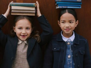 Duas meninas equilibram livros sobre a cabeça diante de uma porta de madeira, em campanha de inverno 2026 da Bonpoint