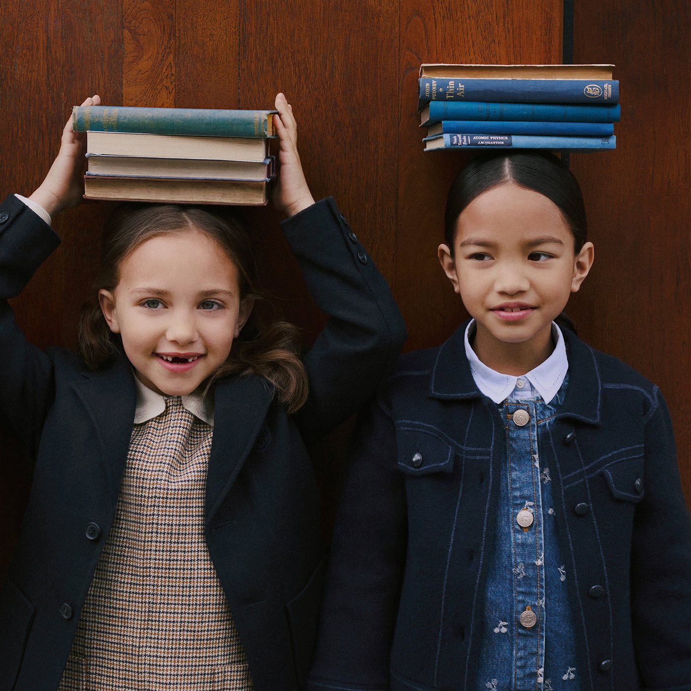 Duas meninas equilibram livros sobre a cabeça diante de uma porta de madeira, em campanha de inverno 2026 da Bonpoint