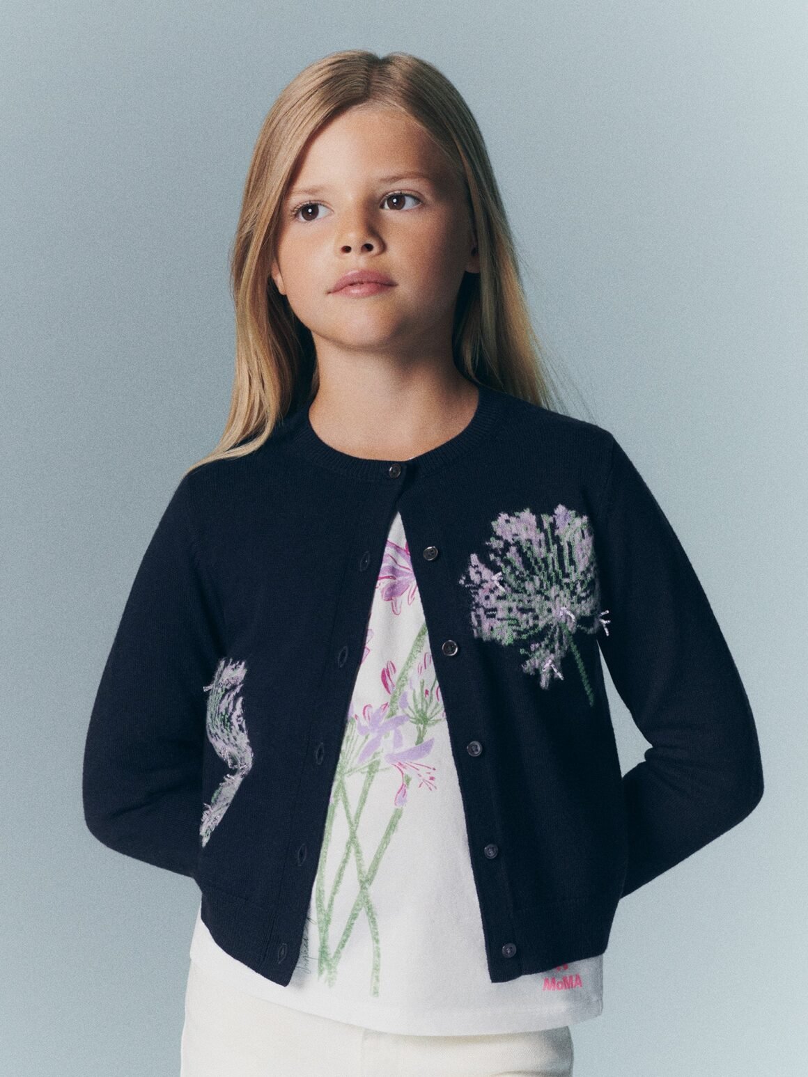 Menina de cardigã azul-marinho bordado com flores lilás sobre blusa estampada, da coleção Bonpoint x MoMA