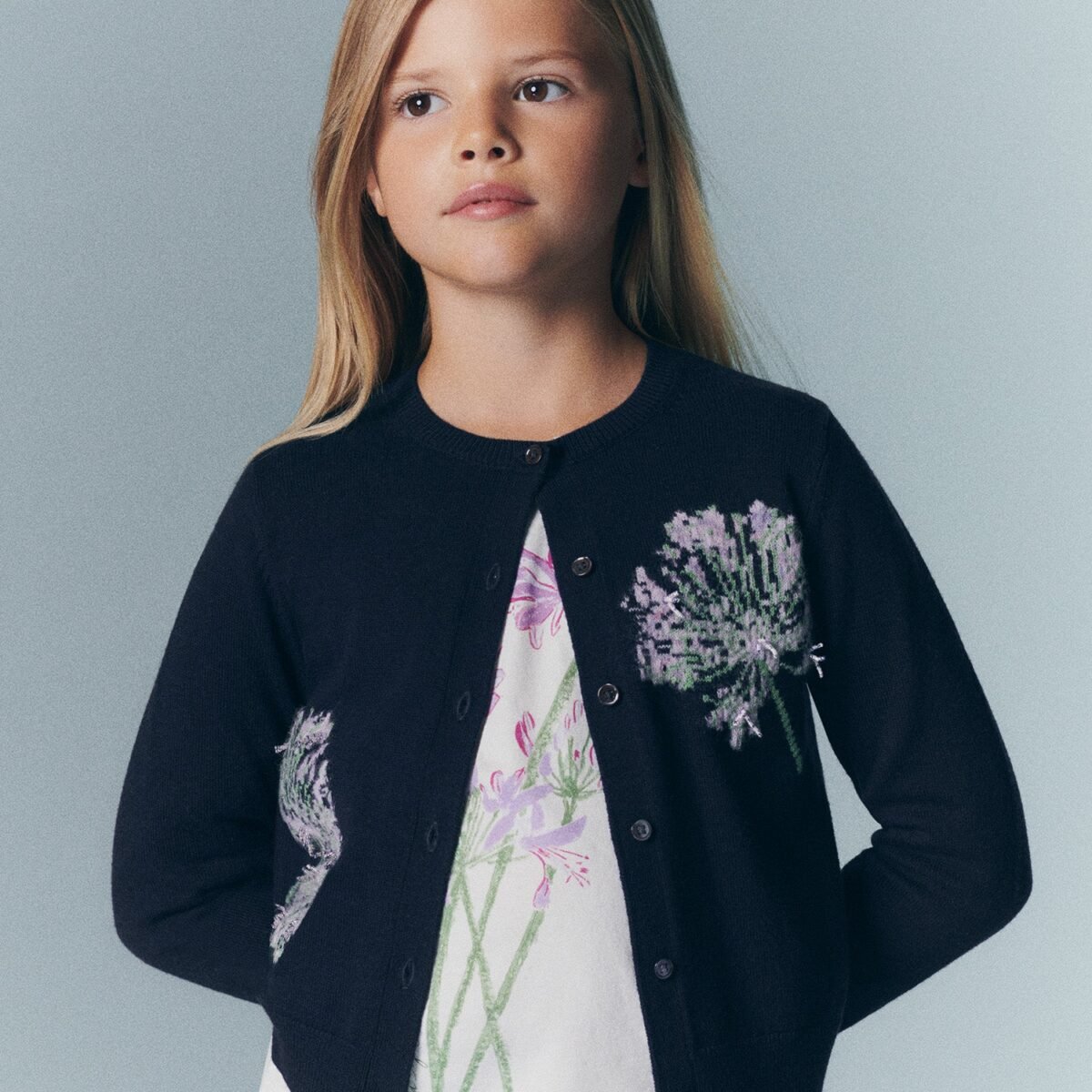 Menina de cardigã azul-marinho bordado com flores lilás sobre blusa estampada, da coleção Bonpoint x MoMA