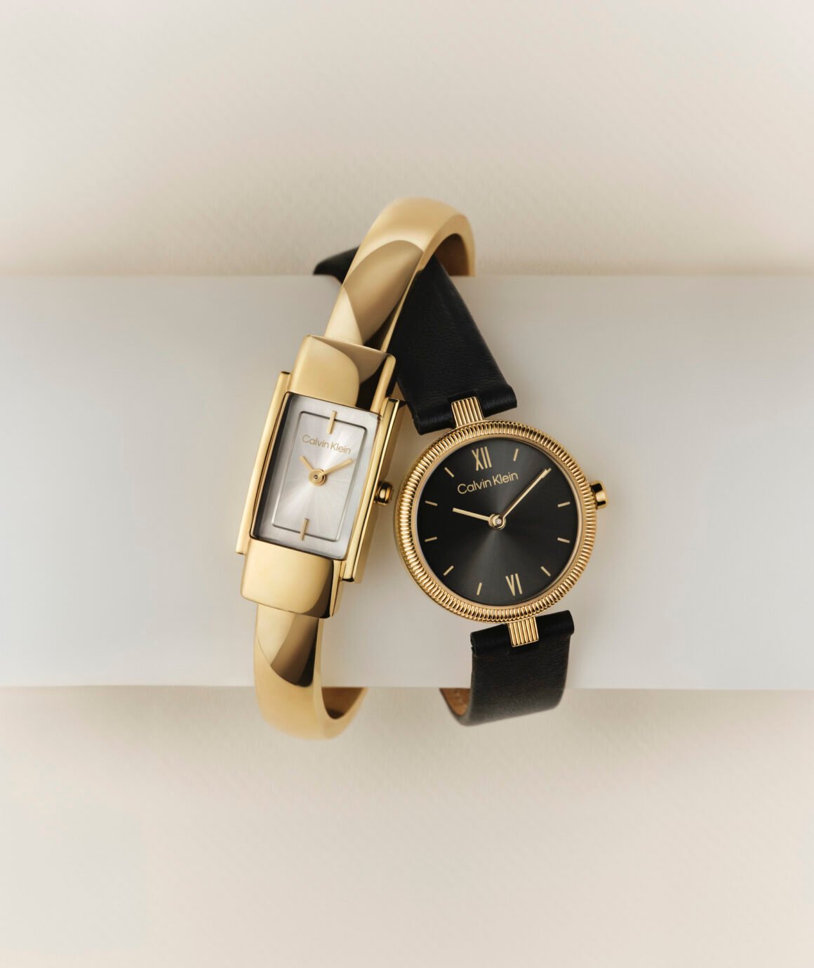 Relógios Calvin Klein dourados, um retangular em bracelete rígido e outro redondo com pulseira de couro preto