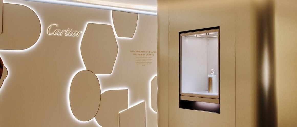 Parede de exposição da Cartier em tom bege, com formas geométricas e um relógio em vitrine.