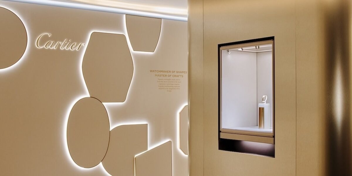 Parede de exposição da Cartier em tom bege, com formas geométricas e um relógio em vitrine.