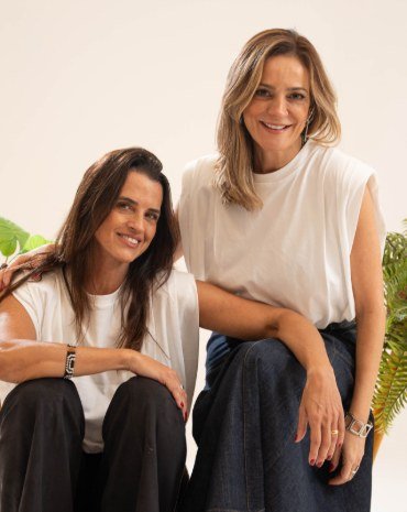 Didi Maakaroun e Vivi Baldeija, criadoras da Casa Rio, de blusas brancas, sorrindo entre plantas