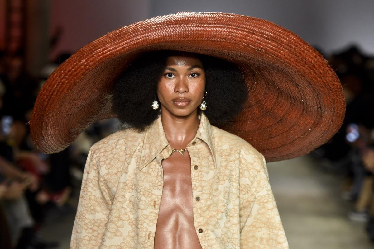 Model wearing an oversized woven hat and a lace-patterned shirt by Dendezeiro on the runway