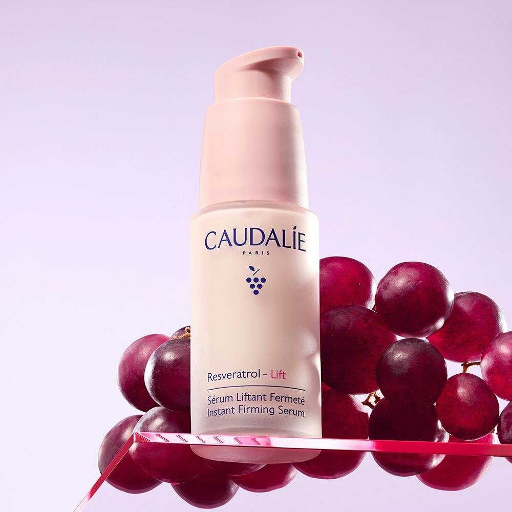 Frasco rosado do sérum Caudalie Resveratrol-Lift apoiado sobre cachos de uva roxa, em fundo lilás