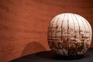 Peça de cerâmica japonesa em forma de esfera, branca com riscos marrons verticais, exposta sobre base escura diante de uma parede de terra avermelhada
