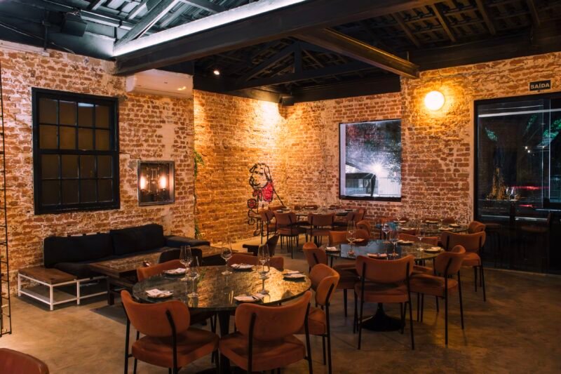 Salão do Clementina Bar de Vinhos, em Pinheiros, com paredes de tijolo aparente e mesas redondas.