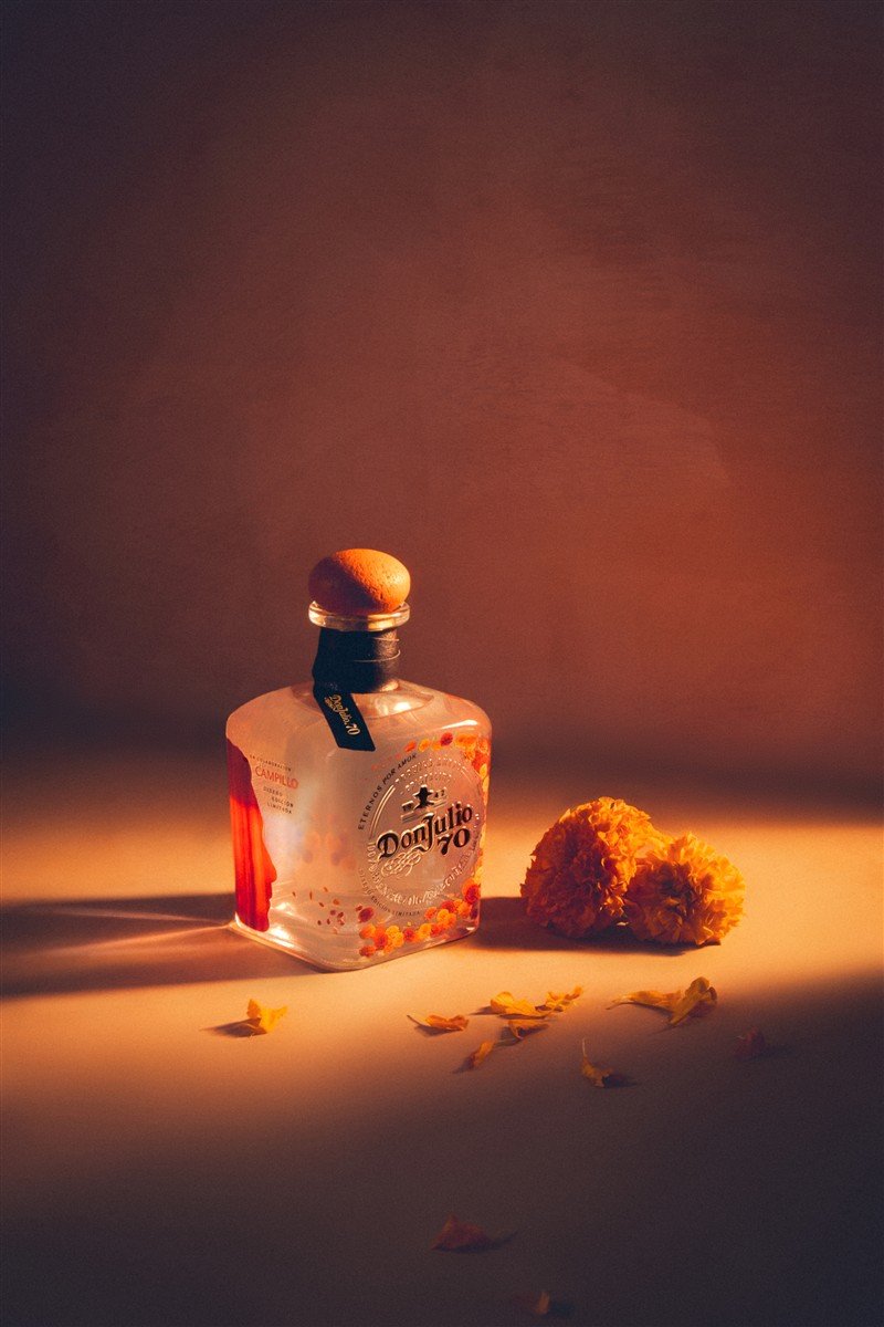 Garrafa transparente de Don Julio 70 decorada com flores alaranjadas ao lado de flores de cempasúchil.