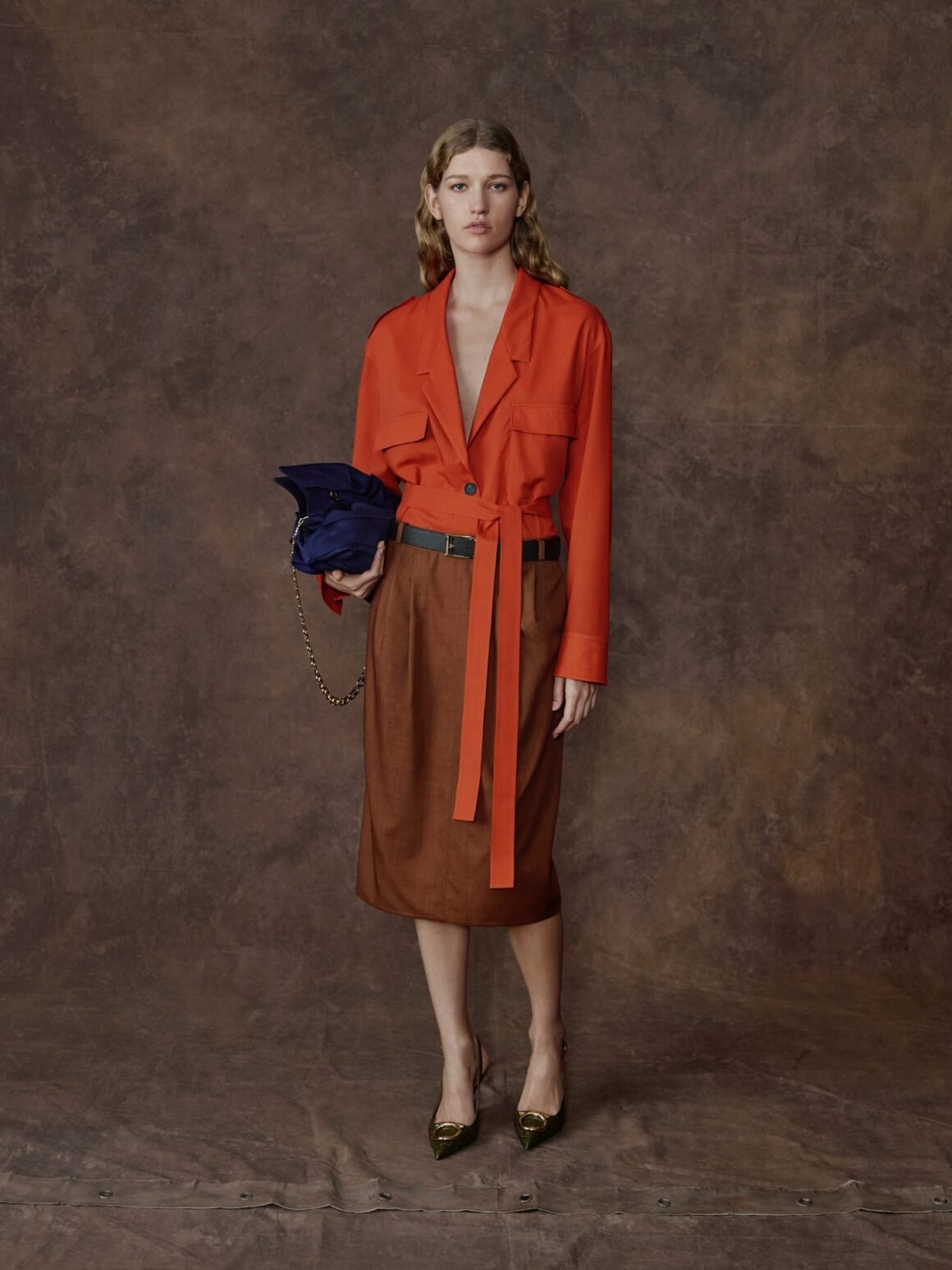 Modelo com jaqueta laranja amarrada na cintura, saia midi marrom e bolsa azul-marinho, em look da Ferragamo