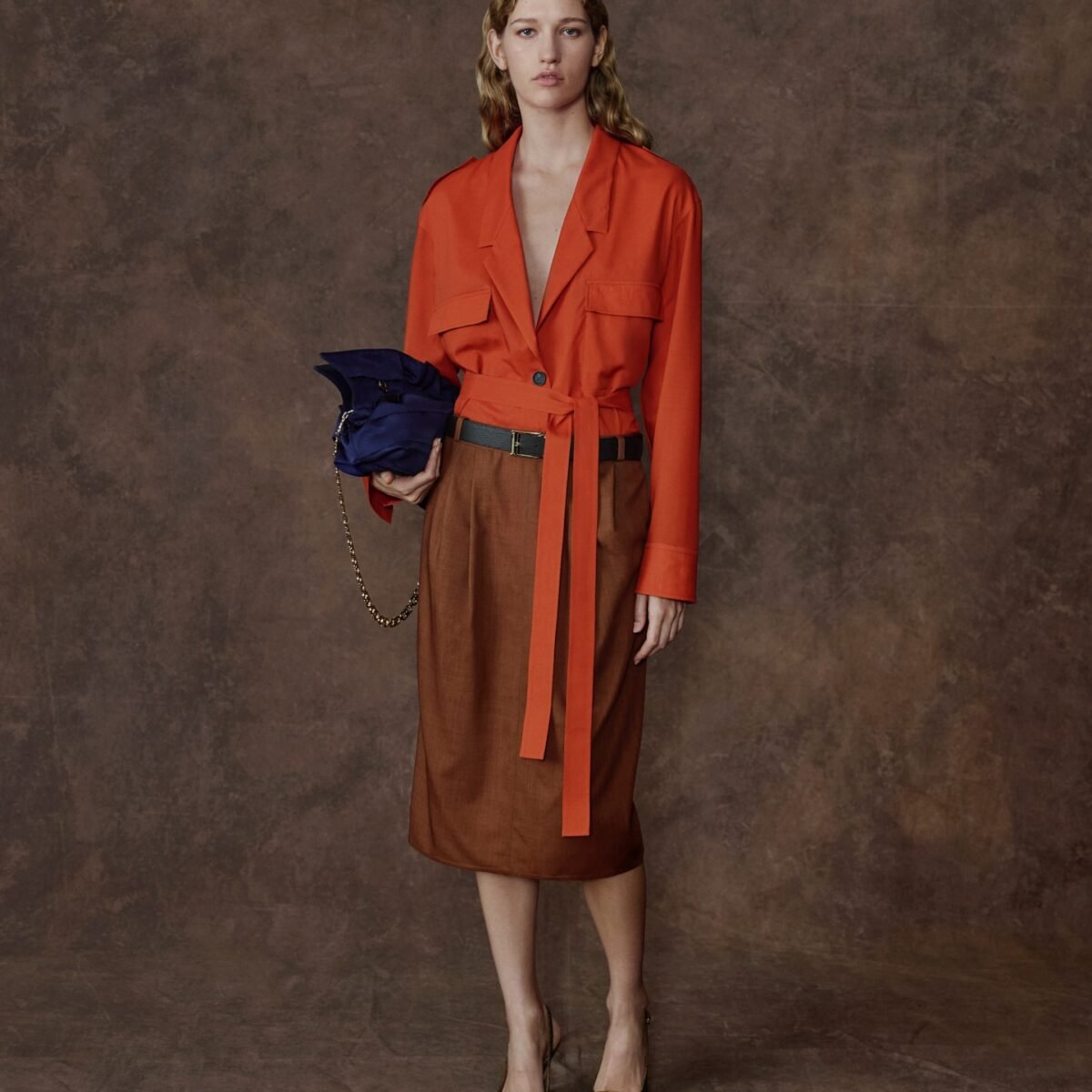 Modelo com jaqueta laranja amarrada na cintura, saia midi marrom e bolsa azul-marinho, em look da Ferragamo