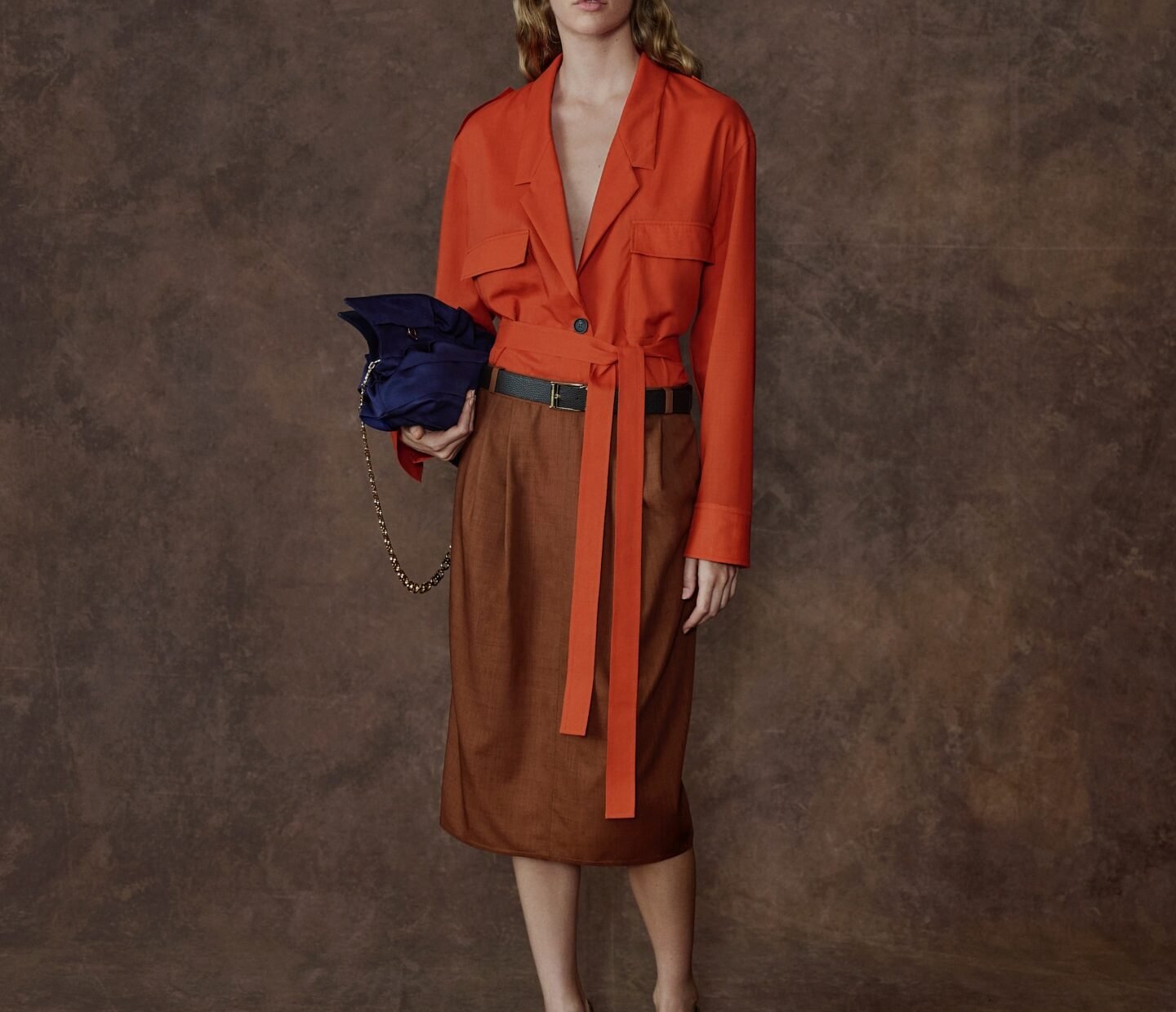 Modelo com jaqueta laranja amarrada na cintura, saia midi marrom e bolsa azul-marinho, em look da Ferragamo