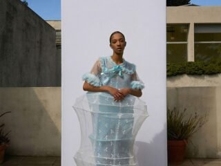 Modelo com vestido de renda azul-clara envolto em estrutura transparente, diante de painel branco