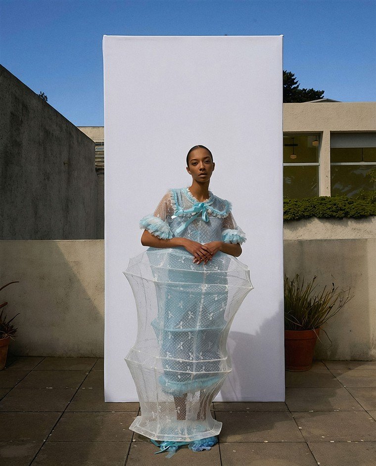Modelo com vestido de renda azul-clara envolto em estrutura transparente, diante de painel branco