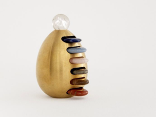 Escultura dourada em forma de ovo com pedras coloridas empilhadas na lateral e uma esfera de cristal no topo, obra da exposição Fluxos II