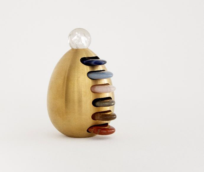 Escultura dourada em forma de ovo com pedras coloridas empilhadas na lateral e uma esfera de cristal no topo, obra da exposição Fluxos II