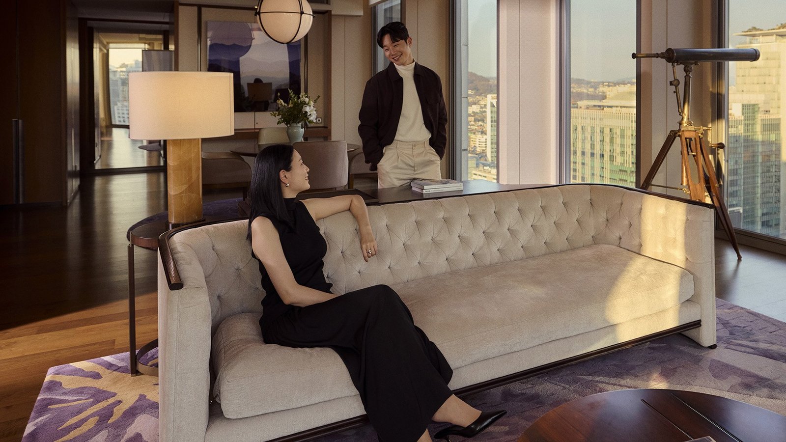 Casal em sala de estar de suíte do Four Seasons Hotel Seoul, com sofá capitonê, luneta e vista para a cidade