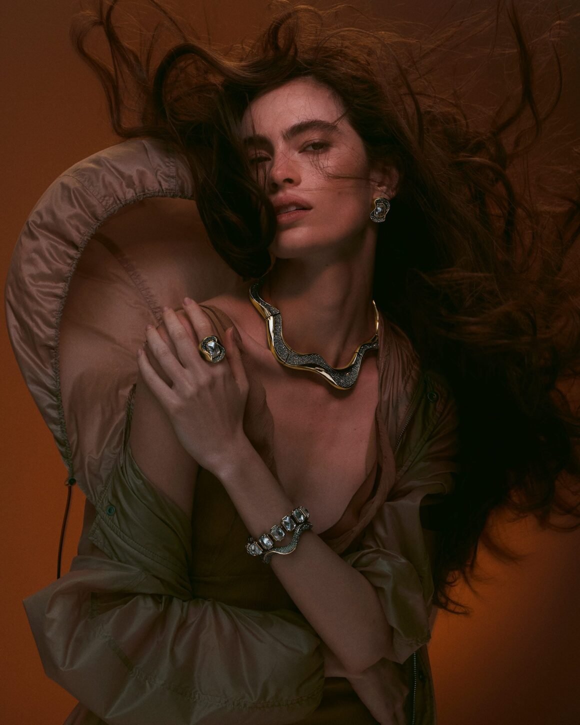 Model with windswept hair in a satin parka, wearing a gold wave-shaped choker, matching earring and ring, and crystal bracelets