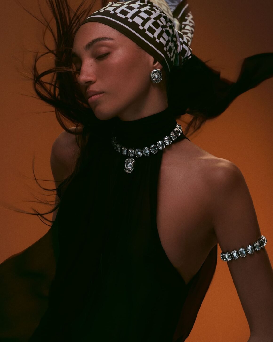 Model with eyes closed and hair in motion, wearing a crystal choker with pendant, a sculptural earring and a crystal armlet