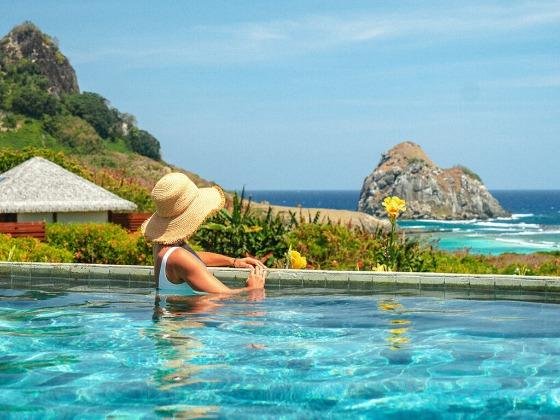 Mulher de chapéu de palha na borda de uma piscina com vista para o mar e o Morro do Pico, em Fernando de Noronha