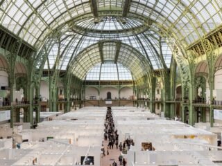 Vista da nave do Grand Palais, com estrutura de ferro e vidro, ocupada por estandes de uma feira de arte.