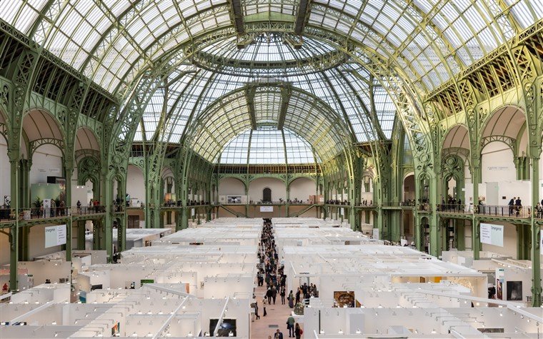 Vista da nave do Grand Palais, com estrutura de ferro e vidro, ocupada por estandes de uma feira de arte.