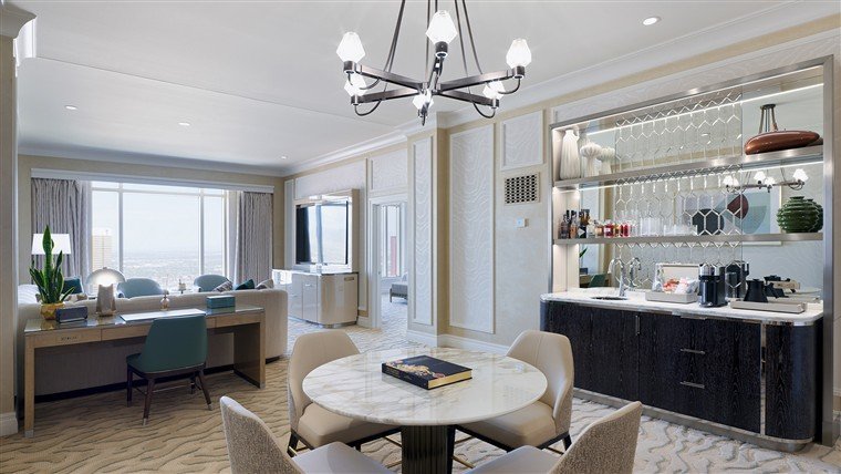 Sala de uma suíte Milano, com mesa redonda de mármore, lustre e bar ao fundo.